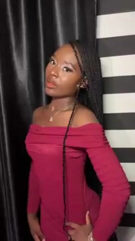A young woman is posing for a photo or video. She has dark skin, black braided hair, and is wearing a maroon off-the-shoulder dress with long sleeves. She is wearing a necklace and a watch. She has makeup on her face. Her right hand is on her hip. The background behind her is half a dark, shiny curtain and half black and white striped wall.