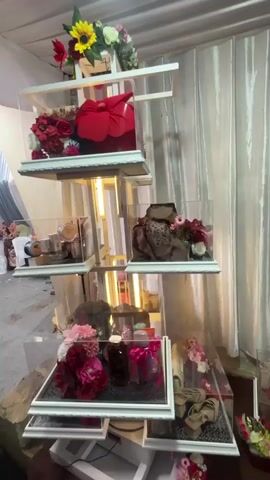 The video shows a tall, multi-tiered display stand filled with gifts, likely for a wedding or engagement. The stand is made of white wood and clear acrylic boxes. Each tier holds several items, including wrapped presents, flowers, shoes, handbags, and other personal accessories. The display is illuminated by lights integrated into the stand. In the background, a white curtain is visible, and to the left, a person in a pink shirt is walking away from the camera. The overall setting appears to be an indoor event space.