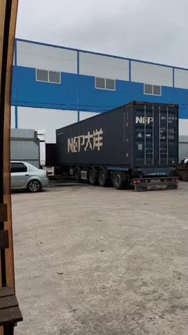 A large blue shipping container on a trailer with multiple wheels is being maneuvered. The container has white text that reads "NCP" and Chinese characters. A silver car is positioned to the left of the container, appearing to be involved in some sort of coordination or assistance. The background consists of a building with blue and white sections, and the ground is paved with a rough surface. The sky is overcast. The video is shot from a slightly elevated angle and focuses on the container and the immediate surroundings.