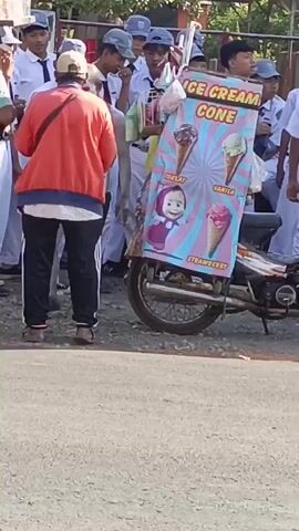 A group of students in white uniforms and some with baseball caps stand near a motorcycle with an ice cream cone advertisement attached to the back. The advertisement features images of four ice cream cones with the flavors Chocolate, Vanilla, and Strawberry listed. A man in an orange jacket and a light-colored hat stands between the students and the motorcycle, appearing to interact with one of the students who is holding a plastic bag. The students are facing towards the motorcycle and the man. The scene takes place outdoors on a paved surface, possibly a street or schoolyard, with trees and a fence visible in the background. The lighting suggests it is daytime, likely sunny.