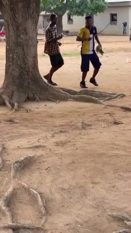 In Tabu, Nigeria, a group of young men is seen running in a circle around a large tree with prominent roots. The ground is dry and sandy dirt.

The men are dressed casually. One wears a bright pink short-sleeved shirt, another a dark tank top with red shorts. A third wears a plaid shirt with dark shorts, and a fourth has on a blue and yellow polo shirt with dark shorts. Another man is in a dark blue shirt and shorts, with a light green fanny pack around his waist. They are wearing various footwear, including sandals and sneakers.

As they run, some of the men are smiling and appear to be enjoying themselves. An audible chant, possibly "Mura" or a similar word, is repeated rhythmically in the background by multiple voices, suggesting a game or group activity. In the background, simple buildings can be seen, further indicating a rural or village setting. The scene takes place during the daytime under clear weather conditions.