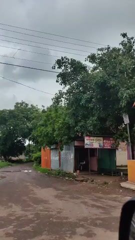 The video begins with a view of a roadside stall or shop, possibly a small eatery, with a flag on a pole in front. A tree with lush foliage is visible behind the stall. The camera then pans to a colorful truck that appears to be selling ice cream, with "Marka Devi" written prominently on its side. The truck has graphics of ice cream cones and other frozen treats. Following this, the video shows the entrance to a building, possibly a temple, with an "Om" symbol above the entrance and signs on either side. Motorcycles are parked in front of the building. The sky is overcast throughout the video.