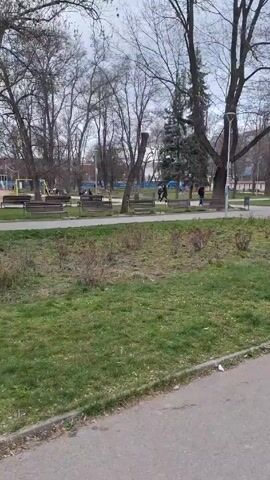 The camera pans across a park. The sky is cloudy. The trees are mostly bare. There is grass, some of which is brown. There are benches and people in the distance. As the camera pans, it focuses on a statue of a person. There is a path leading up to the statue.