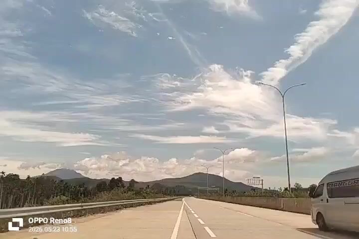 The video shows a view from inside a moving vehicle on a highway. The road is straight and wide, with lane markings visible. There's a white van in the adjacent lane, moving in the same direction. On the side of the road, there are barriers, trees, and hills in the background. The sky is blue with scattered clouds, and there are streetlights along the highway. The timestamp indicates the video was taken on May 23, 2026, at 10:55, and the camera used was an OPPO Reno8.