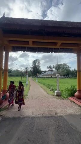 The video shows a brick path leading from under an ornately decorated archway to a white building surrounded by green grass and trees. The archway has yellow posts and a dark red tiled roof. String lights are draped across the archway. Two women in colorful patterned dresses walk from left to right in the foreground. The path is lined with decorative white pillars with curved lampposts. The sky is overcast with scattered clouds.