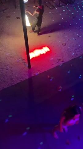 The video shows a person performing a fire show in an outdoor setting. A performer in dark clothing manipulates two flaming torches. The ground is a sandy or stony surface speckled with small green lights.  Several tables and seating areas are visible in the background, suggesting a patio or lounge.  A person wearing a white or light-colored head covering and top is standing near the edge of the frame, looking at the fire performer and occasionally filming with their phone. The scene is dimly lit, with red and purple lights illuminating the foreground.  Music is playing in the background.