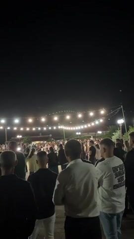 The video captures an outdoor gathering at night. Numerous people are present, some dancing in a circle while others stand around. String lights are strung across the area, illuminating the scene. The foreground features the backs of several men facing the crowd. The overall atmosphere appears to be festive.