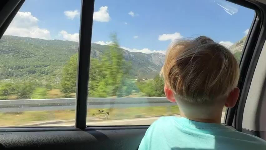 The video shows the back of a child's head with blonde hair looking out the window of a car. The child is wearing a light blue t-shirt. Through the car window, a blurred green landscape with trees and mountains is visible under a bright blue sky with white clouds. The car appears to be moving, as indicated by the motion blur of the scenery. The window has water droplets or dirt specks on it, and a black trim frame is visible. A white guardrail is seen along the side of the road in the distance.