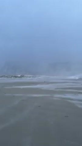 The video shows a beach on a cloudy day. The sand is gray and wet, and the sky is a dull gray-blue. There are waves breaking on the shore, and the water appears rough. A misty haze obscures the view of land on either side of the beach. There are some footprints on the sand in the foreground. The weather is dreary.