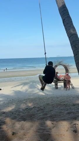 A person is swinging on a rope attached to a palm tree on a sandy beach. The person is wearing a black long-sleeved shirt and yellow shorts, with goggles on their forehead. They are holding onto the rope with both hands and their legs are bent. In the background, there is a calm ocean, a beach with footprints, and a decorative nest-like structure with two people sitting in it. One person is wearing a pink bikini and the other is wearing a black swimsuit. The sky is clear and blue.