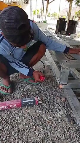A person wearing a blue shirt, black baseball cap, and flip-flops is welding metal pieces in an outdoor environment. The person is crouching and holding a welding torch, which is emitting sparks and bright light as it joins two metal beams. A red and black box labeled "NIKKO STEEL RD-460 WELDING ELECTRODES" is lying on the gravelly ground in front of the person. In the background, there is a yellow water tank, some industrial structures, and two large black speakers. The ground is covered with small stones and gravel. The welding process is ongoing.