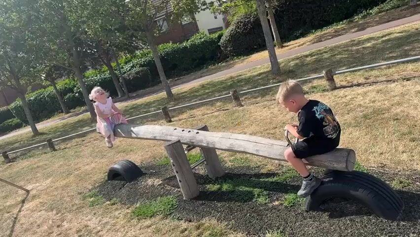 The video shows two young children on a seesaw in a park on a sunny day. A boy with blond hair wearing a black t-shirt and shorts is on one end of the seesaw, and a girl with blond hair wearing a light-colored dress is on the other. They are both facing away from the camera and appear to be actively playing. The seesaw is made of wood and rests on two wooden supports. Tires are placed on the ground beneath each end of the seesaw. The park has dry, yellowish grass and trees in the background, with a paved path and some houses visible further in the distance. The overall environment suggests a warm, outdoor setting.