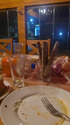 The video shows a table setting after a meal, with dirty plates and glasses. There are also some decorative elements like lights and flowers. The scene appears to be indoors, possibly a restaurant or a home, with a window in the background showing a night view with some lights. In the later part of the video, a man's face is visible in a close-up shot. He is looking upwards and speaking, but his words are not fully discernible in the context provided. The overall impression is a relaxed, post-meal setting.