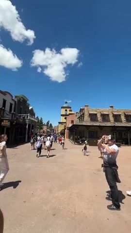 The video shows a street scene in what appears to be a theme park with a Western theme. There are people walking around, and the buildings are designed to look like they are from the Old West. The sky is blue with some white clouds. One building is marked "Bath House 1859". People are dressed in a variety of casual clothing. A man in the foreground is taking photos with his phone.