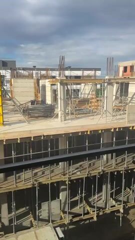 The video shows a construction site with a multi-story building under construction. The building has concrete pillars and floors, with scaffolding and construction materials such as pipes and wooden planks. The sky is cloudy. In the background, there is another building, partially constructed, with brick walls and windows. A crane is visible on top of this background building. The foreground shows a balcony railing, suggesting the video is filmed from a higher vantage point. The scene is static, with no visible people or actions.