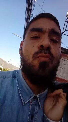 A man with a beard wearing a blue denim shirt is talking to the camera. He is outside during the day and it looks like a clear day. He has a jacket slung over his shoulder. There are wires above him. Behind him there is a white building, and there is a view of mountains in the distance.