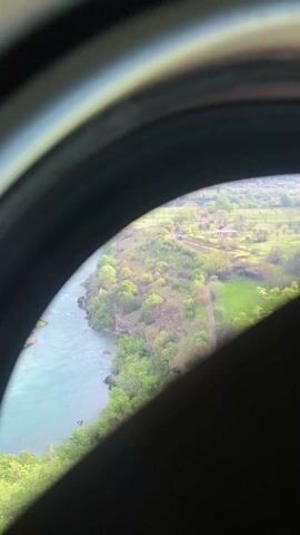 The video shows a view of a river and landscape seen through a rounded frame or tunnel. The river appears to be light blue and runs along the left side of the frame. On the right side, there is a green landscape with trees, grass, and a few houses or buildings in the distance. The view is slightly obscured by a dark, solid shape in the lower right portion of the frame. The video appears to have been taken from an elevated position, such as from a building or a moving vehicle. The overall setting seems to be rural, with a focus on the natural environment.