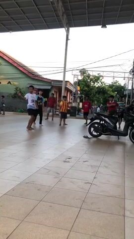 The video shows a group of young people playing soccer under a covered area with a tiled floor. In the foreground, two young males are actively engaged in the game. One, wearing a maroon t-shirt and black shorts, is dribbling a soccer ball and appears to be trying to maneuver around the other player. The second player, in a white t-shirt with a graphic and black shorts, is in a defensive stance, anticipating the ball. A third male, wearing a yellow and red striped shirt, stands to the right, observing the game. Further in the background, other individuals are visible, including a person in a red shirt, and a motorcycle is parked on the right side of the frame. The environment suggests an outdoor recreational space, possibly a schoolyard or community area, with buildings and trees visible in the background. The lighting indicates it is daytime, with sunlight casting shadows on the tiled floor. The action captured is dynamic, with one player mid-action and another appearing to slide or fall while attempting to play the ball. Scattered on the floor are several pairs of flip-flops and some plastic cups.