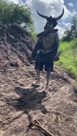 Here is a description of the video:
A man painted gray and wearing a horned headdress gives a piggyback ride to a boy with a backpack, as they climb a rocky hillside covered in vegetation. The man wears black shorts and white and red sneakers. The boy wears long pants and a white shirt.  The ground is covered with rock and vegetation. A shovel lies on the ground. The sky is partly cloudy.
