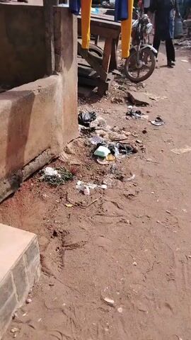 The video depicts an outdoor market or street stall area in Awka Etiti, Nigeria, during the daytime under sunny weather. The ground is a mix of reddish-brown dirt and mud, heavily littered with various types of trash, including plastic bags, food containers, and other debris.

On the left side of the frame, there is a low concrete or cement structure. Clothes, specifically a pair of yellow pants and a blue garment, are hanging to dry on a wooden post next to this structure.

In the mid-ground and background, several wooden tables, likely market stalls, are visible. One table has a blue plastic sheet covering white contents, possibly grains or other foodstuffs. Another table is empty, showing its wooden plank surface. A motorcycle wheel is partially visible near the tables.

In the far background, under what appears to be a covered market area, a person wearing blue pants and a dark top is seen walking away. Other blurry figures are also discernible, suggesting activity in the wider market. The overall scene portrays a typical informal market environment, characterized by its functional setup and the presence of litter.
