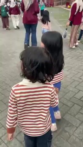 The video shows a bustling outdoor scene. Initially, two young girls in striped shirts and blue bottoms stand on a brick walkway surrounded by a crowd of people. The scene then pans to show a busy marketplace filled with people and stalls. Finally, the camera focuses on a unique building with a red exterior and a multi-tiered green roof, reminiscent of pagoda architecture, with a black cube structure mimicking the Kaaba attached to one side. People are milling about in front of the building. The sky is overcast.