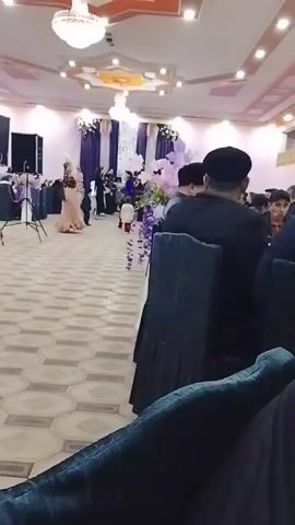 The video shows the inside of a large, well-lit banquet hall decorated for a wedding or celebration. The hall features a patterned tile floor, a decorative ceiling with chandeliers, and purple and white floral arrangements. Several people are present, some seated at tables covered with white cloths and others standing near a decorated stage area. Children, dressed in formal attire, are running down the aisle between the tables. The camera is positioned from a seated perspective, focusing on the people and activities in the hall. The general atmosphere suggests a festive and lively occasion.