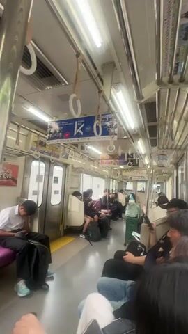 The interior of a mostly empty subway car is seen in this video. The subway car is brightly lit by fluorescent lights. Several people are sitting on the seats, most are looking down at their phones or sleeping. Many white loop handrails with tan straps hang from the ceiling. A poster ad hangs above the handrails. The walls are white with silver trim. A set of closed doors with a yellow stripe are seen on the left of the frame.