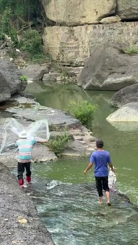 Two individuals are standing near a body of water. The person on the left is wearing a hat, a striped shirt, black pants, and red boots. They are holding a net, which is spread out and appears to be in the water. The person on the right is wearing a blue shirt, dark pants, and a baseball cap. They are holding a white bag in their right hand. They are both facing away from the camera and towards the water. The environment appears to be a rocky, natural setting with a river or stream flowing through it. There are large rocks and vegetation visible. The water is a greenish color.