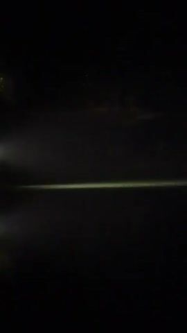 A dark video shows a light source illuminating a foggy or misty area over water. A structure with lights on appears in the distance.