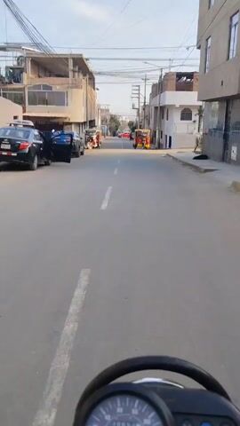 The point-of-view video shows a person riding a motorcycle down a paved street in a residential area. The street has a center dividing line, with parked cars on either side. Several auto rickshaws are also parked along the sides of the street. The buildings on either side are two to three stories tall, with some under construction. Power lines run above the street. In the distance, a person is walking across the street. The motorcycle's handlebars and speedometer are visible in the foreground. The speedometer shows a speed of around 30 km/h. The day is sunny.