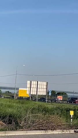 The video begins by showing a clear blue sky with some clouds. Below the sky is a body of water with a distant shoreline lined with trees. In the foreground, there is a road with several cars driving on it. The camera pans to the right, revealing a white SUV parked on the side of the road. The SUV has a license plate that reads "M475TH164". The video continues to pan right, showing more cars driving on the road and a street light. The environment appears to be an outdoor scene with vegetation and a road.