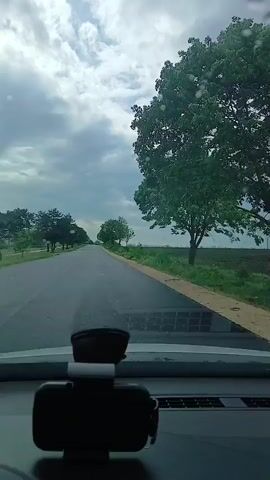 The video shows a car driving on a two-lane road. The sky is cloudy and there are trees lining the road. There is a phone holder on the dashboard. Raindrops can be seen on the windshield.  Music is playing in the background.
