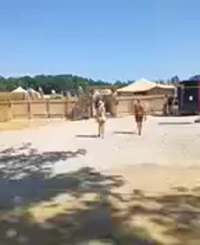 Two people walk across a dusty lot in front of a fenced-off area with tents and a large black vehicle. The sky is clear and blue. The ground is light brown and covered in small stones. Trees and hills are visible in the distance. The person on the left is wearing a light-colored outfit, and the person on the right is wearing a dark outfit. The vehicle appears to be a truck or bus. There are several tents or canopy structures visible.