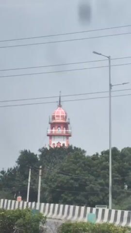 This is a vertical video shot from a moving vehicle, likely a car or truck. The camera is focused on a tall, white and pink tower with a red dome, which is partially obscured by trees. The tower appears to be a significant structure, possibly a landmark or a building with a unique architectural design. In the foreground, there is a rusty, orange-colored object, which is also part of the vehicle the camera is in. The sky is overcast and gray, suggesting a cloudy day. Power lines are visible crisscrossing the sky above the tower. In the lower part of the frame, glimpses of other vehicles and road infrastructure can be seen, confirming the perspective of a moving vehicle on a road.