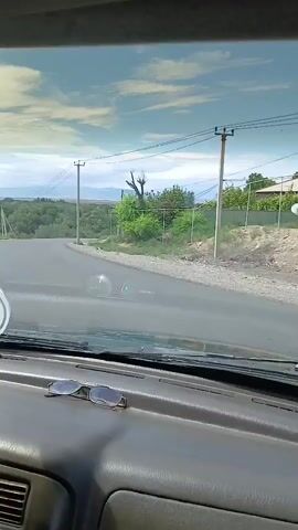 The video shows a view from inside a car, looking out the windshield. The car is driving on a paved road that curves to the right. On either side of the road, there are trees and vegetation. In the distance, there are mountains under a partly cloudy sky. There are power lines running along the road, supported by utility poles. The dashboard of the car is visible in the foreground, with a pair of sunglasses resting on it. The lighting suggests it is daytime.