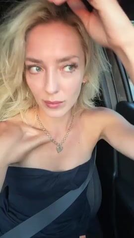 This media file shows a close-up, waist-up shot of a young woman with blonde hair and fair skin. She is seated in a car, indicated by the presence of a seatbelt and the car's interior visible in the background. The woman is wearing a black strapless dress and a gold necklace with a heart-shaped pendant. She is touching her hair and looking towards the camera with a neutral expression. The lighting suggests it is daytime, possibly late afternoon, with natural light illuminating her face. The background outside the car window shows greenery, suggesting a somewhat rural or suburban setting. The scene appears to be filmed in Beit Hanan, Israel. There is no visible text in the media file. The woman's actions and the close-up framing create an intimate and personal feel to the shot.