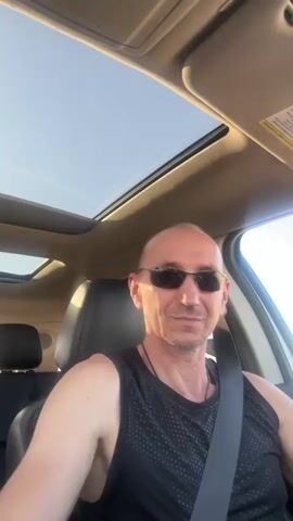 A man in sunglasses and a black tank top is sitting in the driver's seat of a car. He is wearing a ring on his left hand. He is smiling and making a heart shape with his thumb and index finger. The sun is shining through the sunroof of the car.