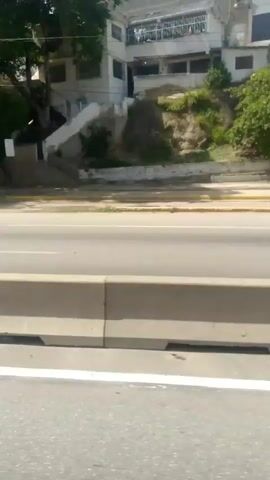 The media file shows a view from a moving vehicle on a highway in Macuto, Venezuela. The scene depicts a highway with multiple lanes separated by concrete barriers. On the side of the highway, there are green trees, vegetation, and some buildings on a hillside in the background. The sky appears bright, suggesting it is daytime, likely with clear or partly cloudy weather.

The primary activity is travel along the highway. Several vehicles, including a black SUV, are visible on the road, indicating ongoing traffic. The perspective is from a passenger inside a vehicle, looking outwards. The environment suggests a somewhat urban or suburban setting with a natural landscape of hills and vegetation adjacent to the man-made infrastructure of the highway. There is no discernible text in the media file. The overall mood is neutral, focused on the journey and the surrounding environment.