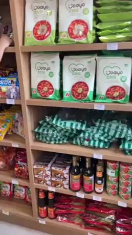 A slow pan of the shelves of a small Korean grocery store reveals the aisles are stocked with an assortment of products. The first shelf is full of packages of "Joayo" Korean chili noodles and other Asian snacks. The next shelf down is stocked with individually wrapped green and white checkerboard-patterned packs. The third shelf contains a variety of Asian sauces and tomato paste cans. A child in a white dress with a red ribbon walks past the shelves on the right side of the aisle. These shelves are stocked with a variety of individually packaged noodles, ramen, and other Asian snack foods.