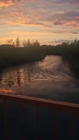 The video shows a river scene at sunset. The camera is positioned on a bridge, looking down the river. The river is calm, with the sunset reflecting in the water. On either side of the river, there are trees and bushes, silhouetted against the colorful sky. The sky is filled with orange, pink, and purple clouds.  The railing of the bridge is visible in the foreground. There are no people visible in the video.