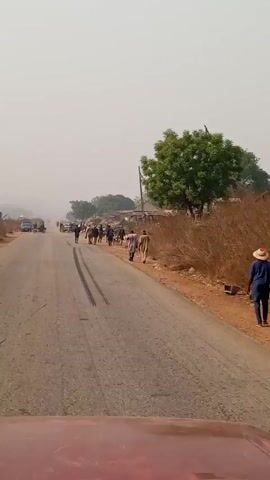 The video shows a scene on a rural road, likely in Africa based on the style of buildings, attire of the people, and the general environment. The camera is mounted inside a moving vehicle, looking down the road. There are several people walking along the roadside, some herding goats. There are also a few parked cars visible. The road is paved but has evident tire marks. The sky is overcast with a hazy quality. The surroundings consist of sparse vegetation and simple structures. The video is shaky, and the focus changes sporadically.
