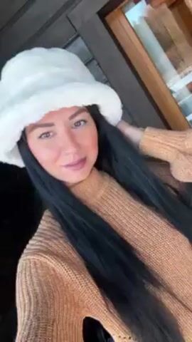 The video shows a woman posing for the camera. She is wearing a tan sweater and a white, furry bucket hat. She has long, dark hair. She is smiling slightly. Behind her is a wooden wall and a window.