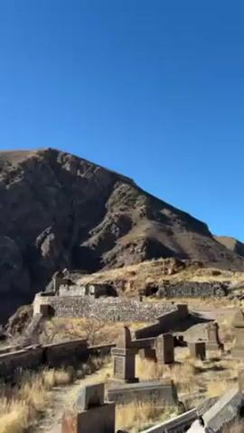 The video pans across a graveyard located on a hillside with a mountain in the background. The graveyard contains stone headstones and walls. Some trees are bare. The sky is clear and blue.