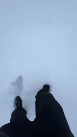 A first-person view of a person walking through deep snow. The person is wearing black pants and dark shoes. They are stepping into the snow, leaving footprints. The snow is white and covers the ground. The wind is blowing.