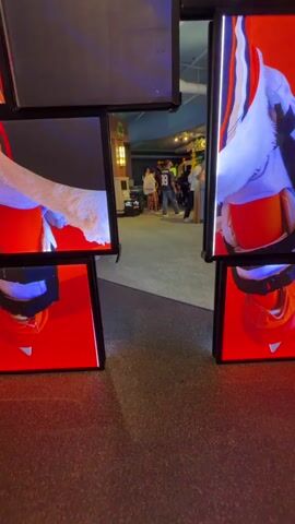 The video shows a view through an entryway that is framed by a mosaic of digital screens displaying parts of a hockey jersey. The jersey is black with orange and white accents and has the name "WILD WING" and the number "93" on it. Several of the screens show close-ups of a furry white appendage that appears to be part of a mascot costume, as well as parts of a white and orange helmet. In the background, through the entryway, several people are visible walking in what appears to be an indoor space. One person in the background is wearing a shirt with the number 99 on it. The floor in the foreground is carpeted with a dark, speckled pattern. The lighting is dim with bright highlights from the digital screens.