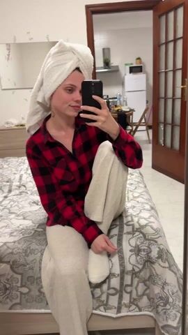 A young woman with a white towel wrapped around her head sits cross-legged on a bed in a bedroom. She is wearing a red and black plaid shirt and cream-colored pants. She holds a black smartphone in her right hand, taking a selfie. A mirror reflects the room behind her, showing a kitchen area with a white refrigerator and a table with various items on it. The bedroom has a dresser and a bed with a patterned grey and white comforter. The woman smiles and adjusts the towel on her head.