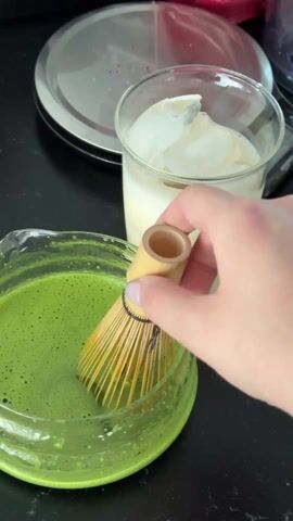 The video shows someone preparing a matcha latte. The scene takes place on a black countertop. To the left is a glass bowl filled with bright green matcha tea, which is speckled with small bubbles.  To the right is a clear glass filled with milk and ice. A bamboo matcha whisk is lifted out of the bowl.  The hand, adorned with silver bracelets and blue nail polish, picks up the bowl of matcha and pours it into the glass with milk and ice.  In the background, a circular metal disc rests on the counter next to a digital scale.
