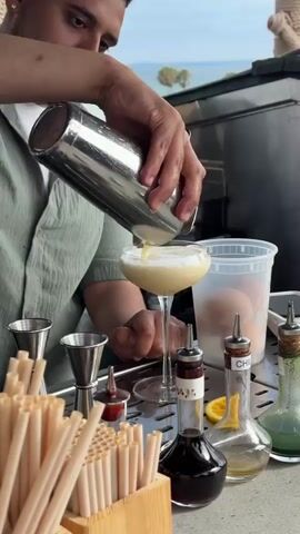 A bartender is pouring a creamy yellow cocktail from a silver shaker into a coupe glass. The bartender is wearing a light green shirt. In the foreground, there is a wooden holder filled with beige straws. Behind the glass, there are two jiggers, a container of eggs, and several bottles of liqueurs or syrups. The setting appears to be outdoors, possibly a bar by the water, with a glimpse of the sky and some greenery in the background.