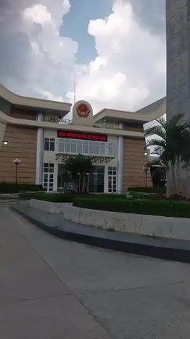 The video captures a view of a border crossing with a large, modern building and a flag flying. The building has signage in a language that appears to be Vietnamese, with the words "CỬA KHẨU QUỐC TẾ MỘC BÀI" visible. There are palm trees and landscaping in front of the building. The camera then pans to the right, showing a large digital billboard and several vehicles, including a white bus. Further panning reveals more vehicles and buildings in the distance, as well as a large arched structure, possibly part of another border control area. Several motorcycles and a red double-decker bus are also visible. The overall environment suggests an international border crossing point with typical infrastructure and traffic. The sky is cloudy.