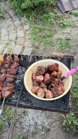 A high-angle shot captures a metal outdoor grill with hot coals. Skewers of meat are cooking on the grill. On the left, a skewer holds chicken wings, and on the right, skewers hold pieces of pork or beef. A metal bowl containing more cooked meat sits on the right side of the grill. The background shows green foliage and a brick-paved area.
